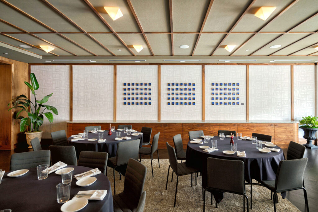 Banquet-style dining room with round tables set with plates and glasses, black tablecloths, and chairs, against wood-paneled walls and plants.