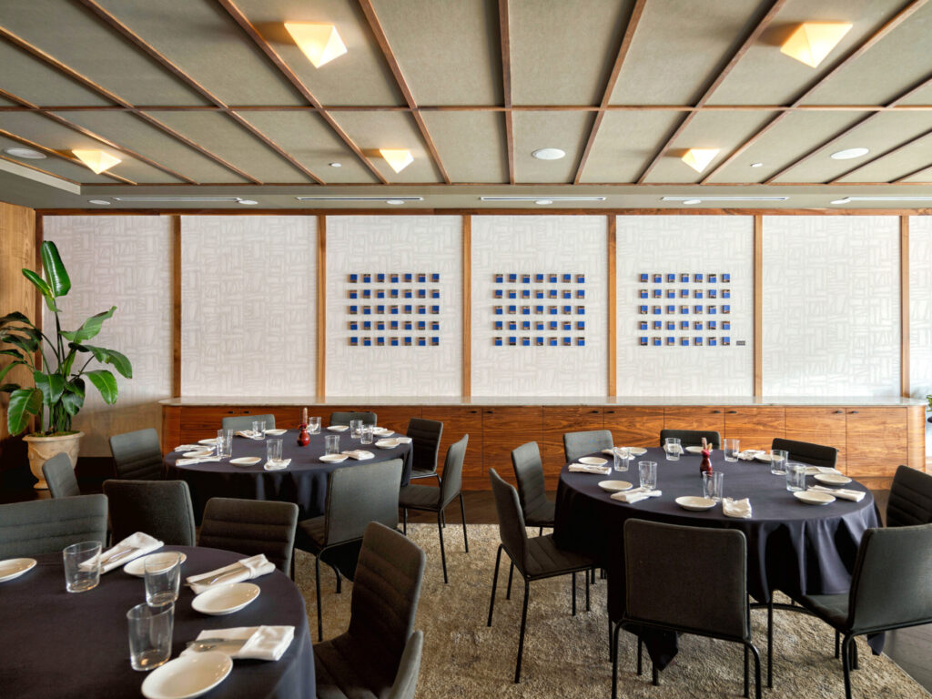Banquet-style dining room with round tables set with plates and glasses, black tablecloths, and chairs, against wood-paneled walls and plants.