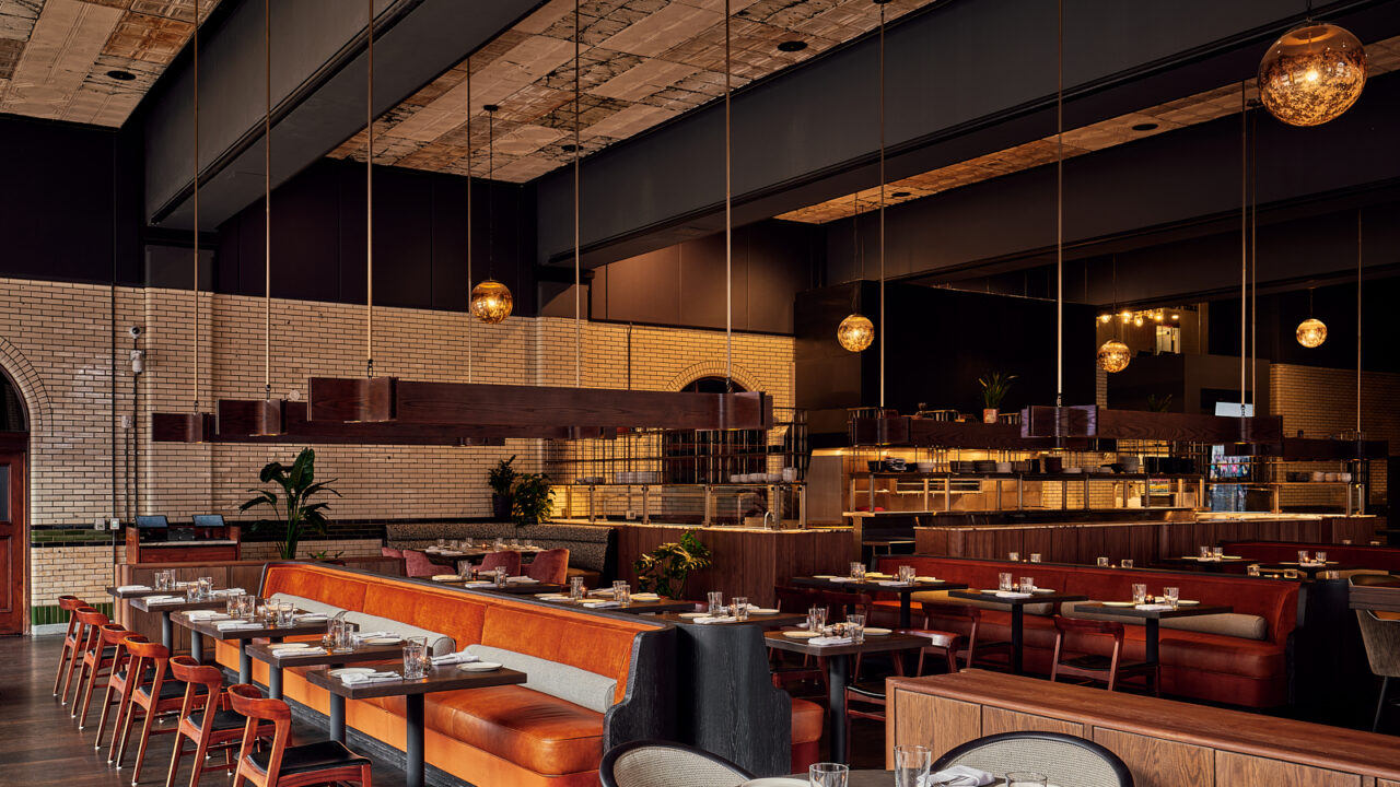 Modern upscale restaurant interior with an orange leather banquette, dark wood tables, and an open kitchen in the background.