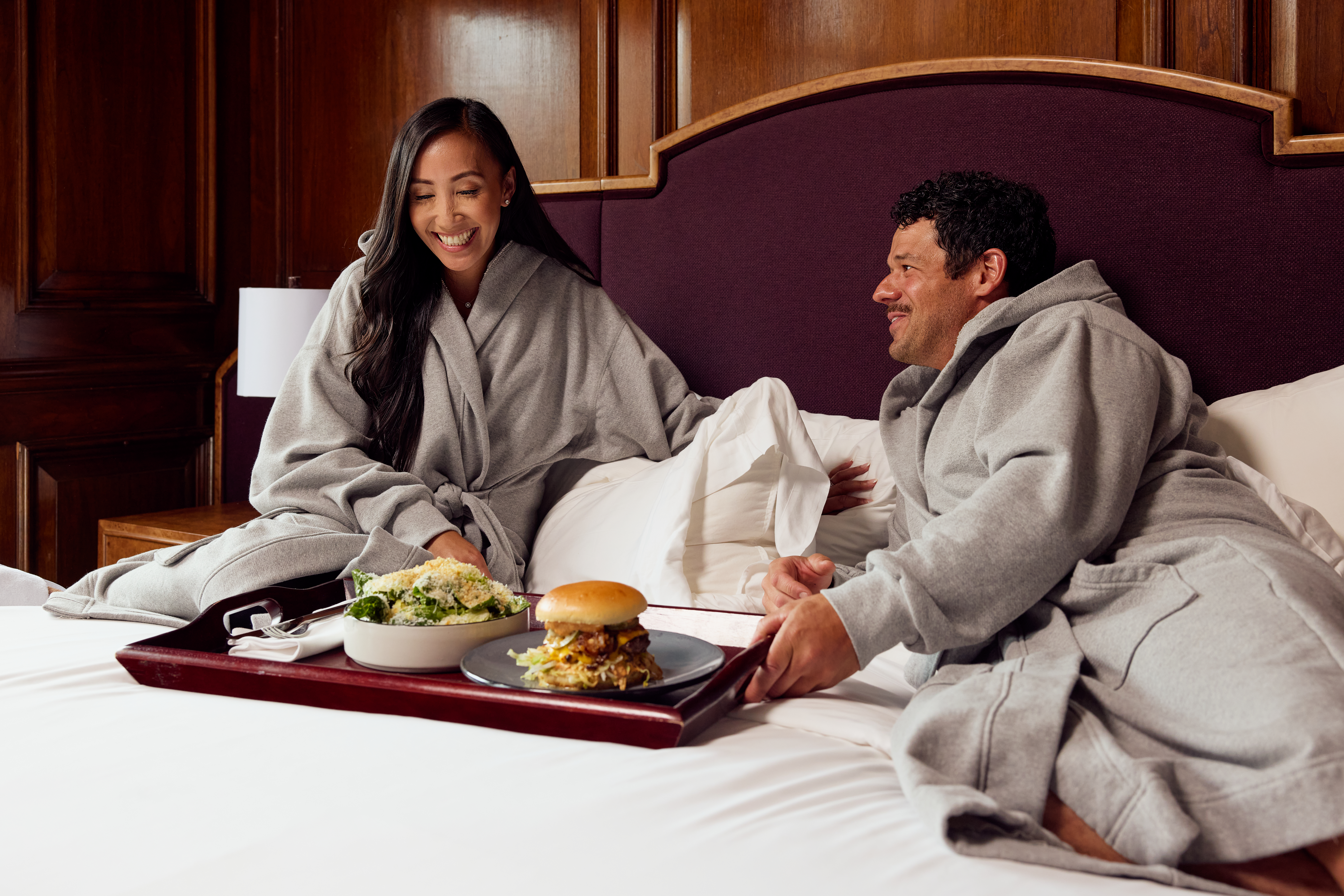 2 people enjoying food on a hotel bed