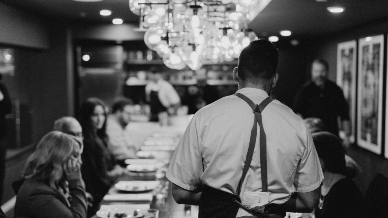 Black and white image of the back of a chef setting a table