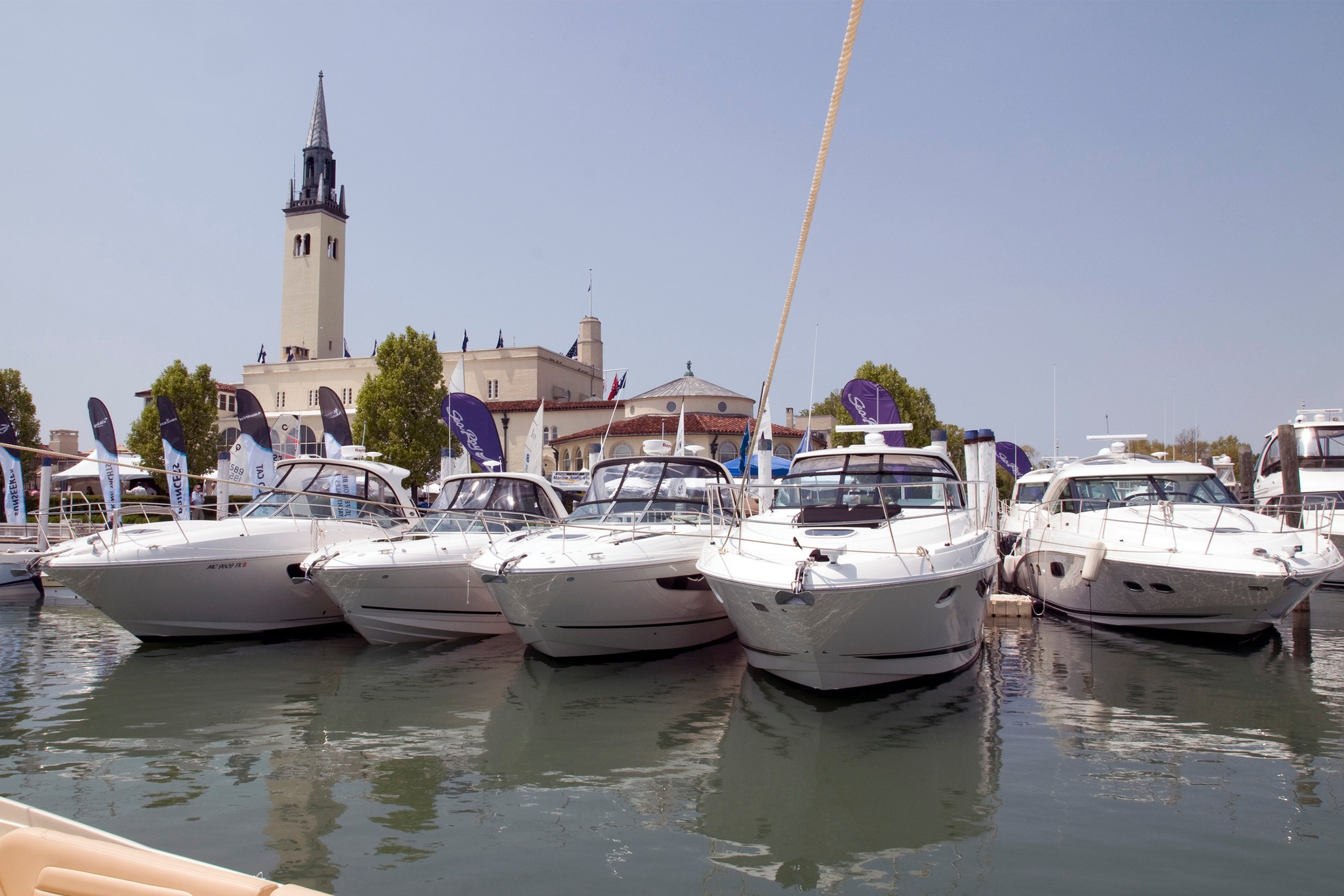 Marina with multiple white motorboats docked, flag banners, and a tall beige clock tower building in the background under a clear blue sky.