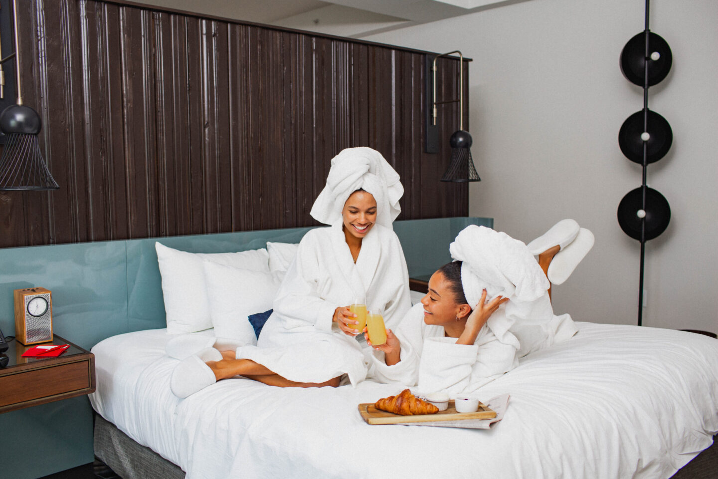 Three women in white bathrobes with towels on their heads sit on a bed, toasting with glasses of orange juice in a hotel spa setting.