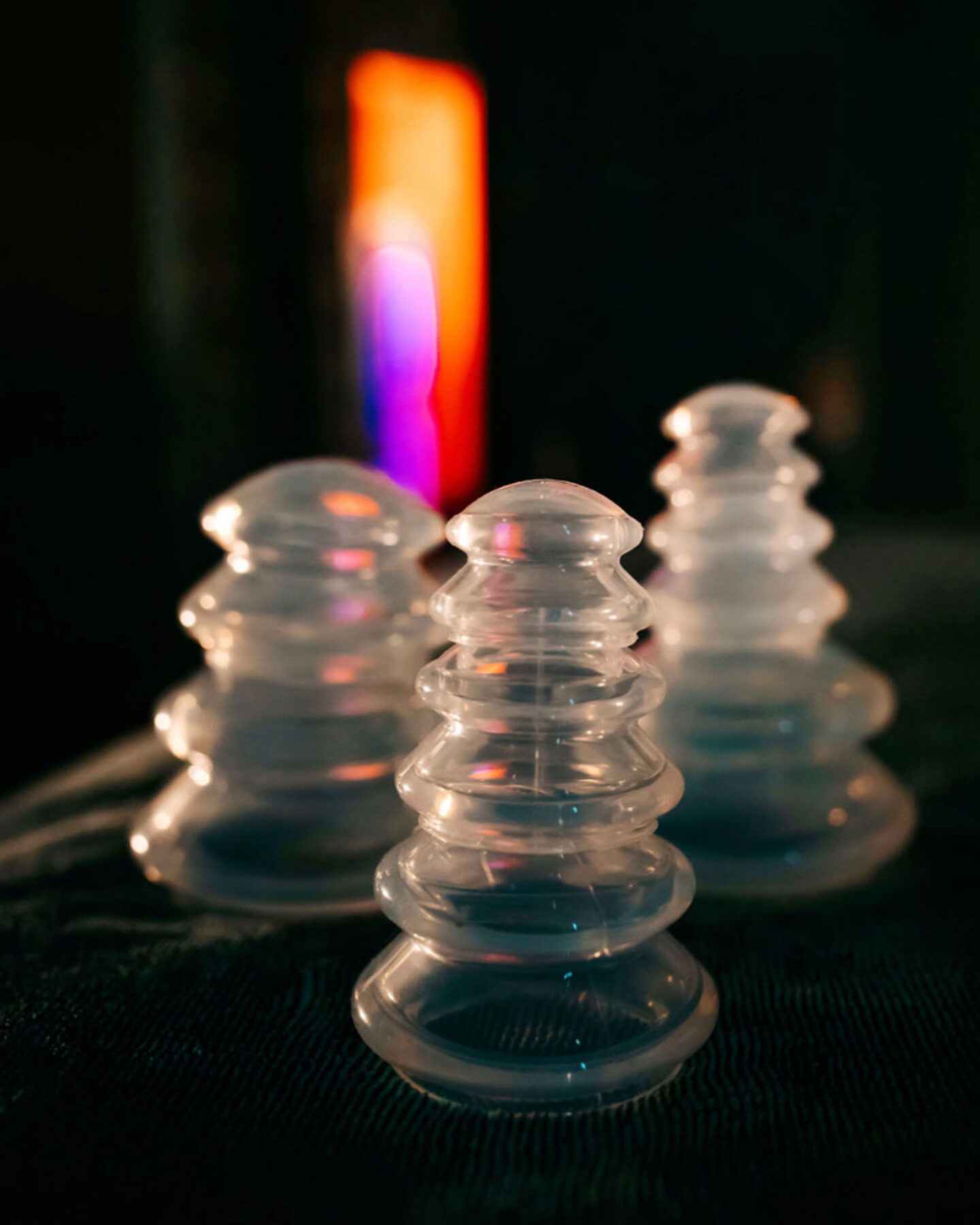 Three translucent glass stackable sculptures arranged on a dark surface with a blurred multicolored light in the background