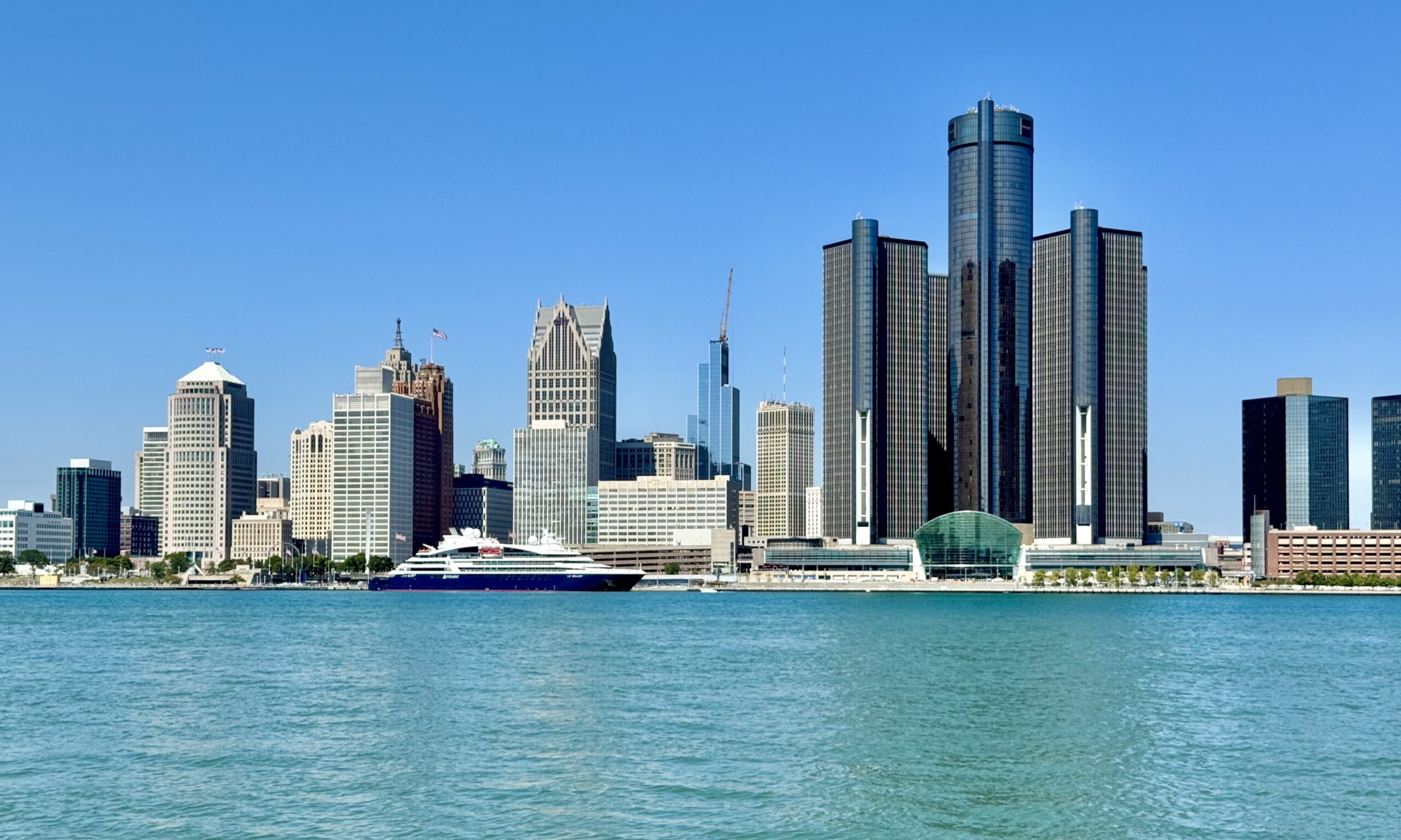 City skyline along a calm blue river with a large white yacht at the waterfront in the foreground.