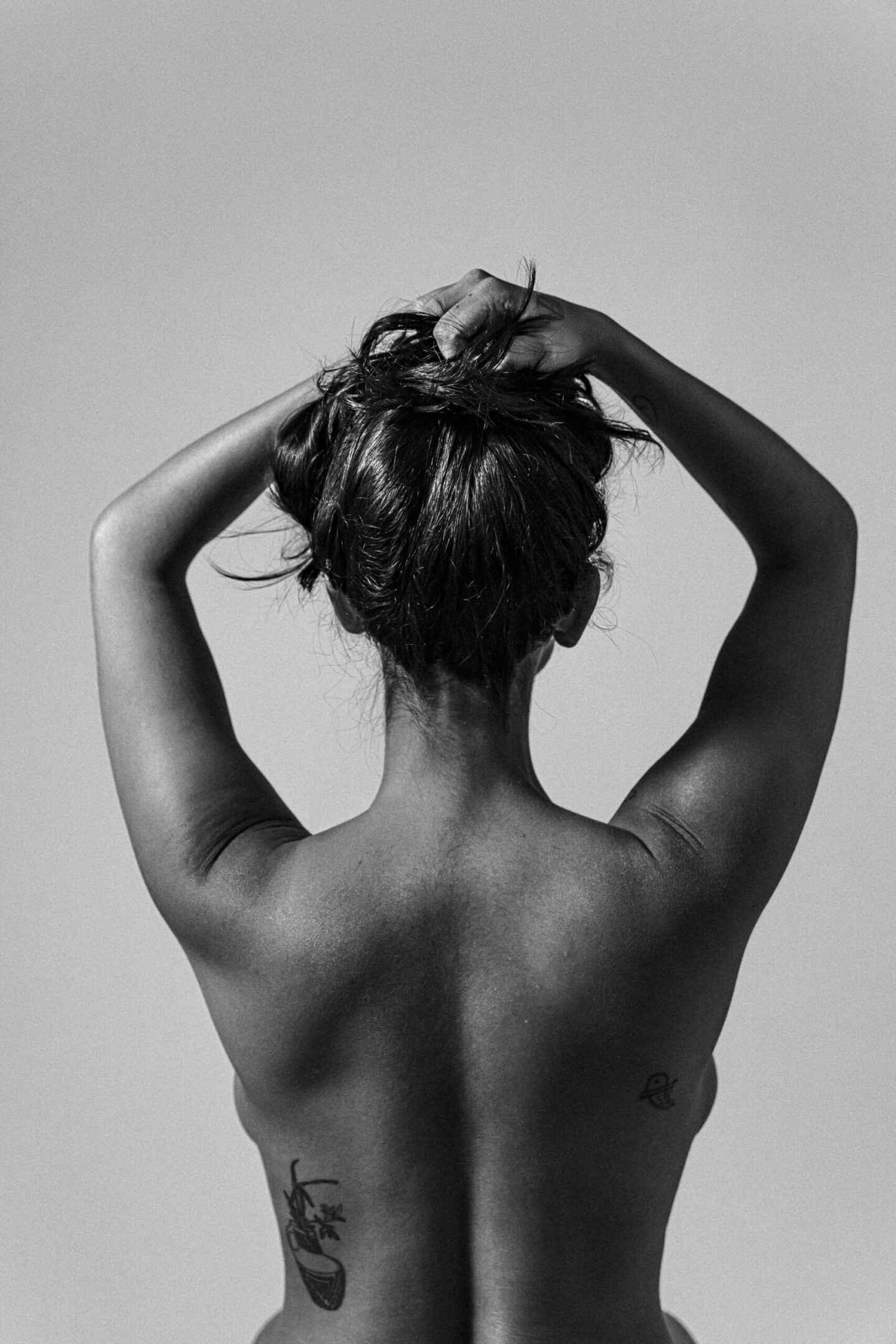 Back view of a topless person tying up their hair, arms raised; visible tattoos on the lower back.
