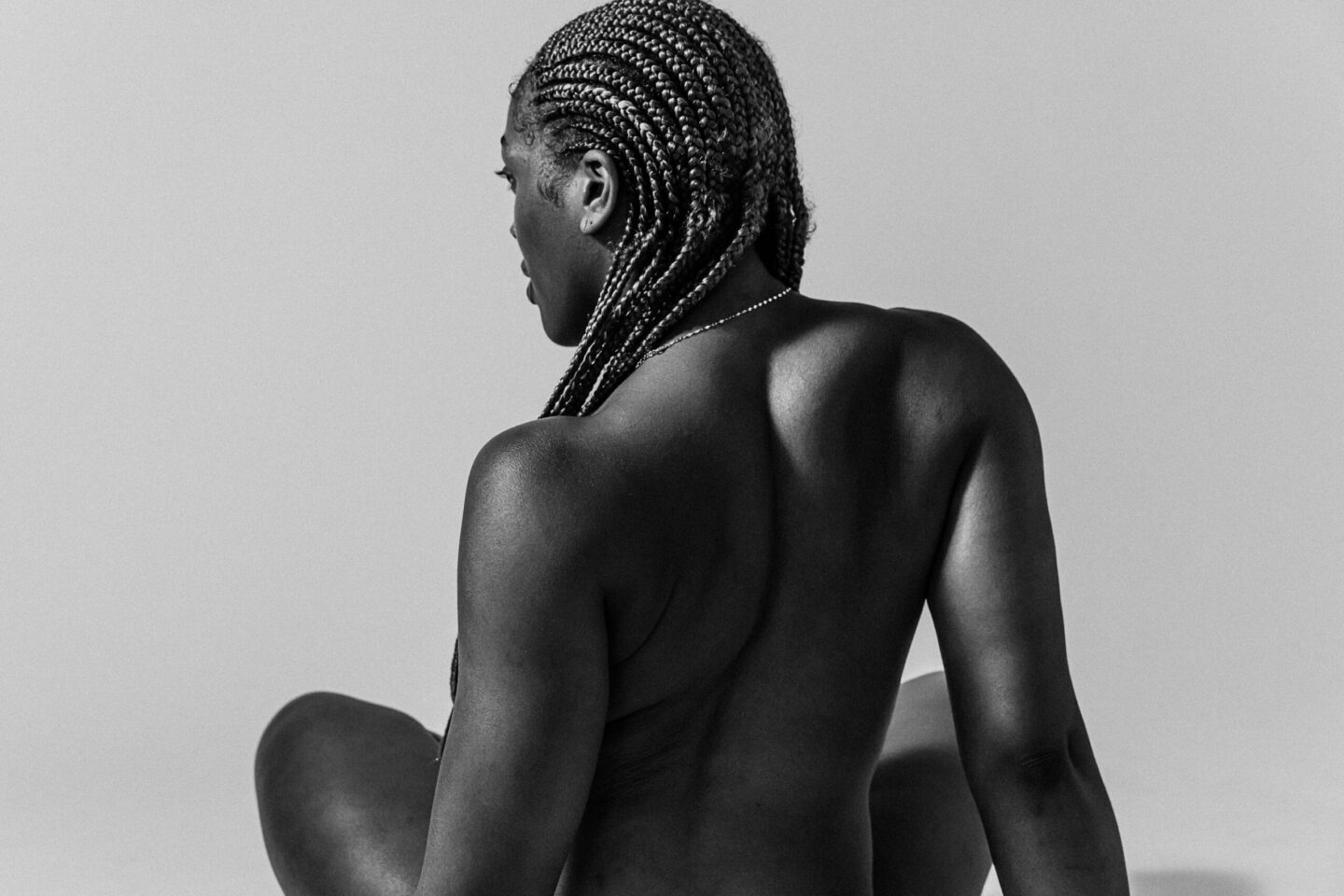 Back view of a topless person with tight cornrow braids and a small necklace, against a plain gray backdrop.