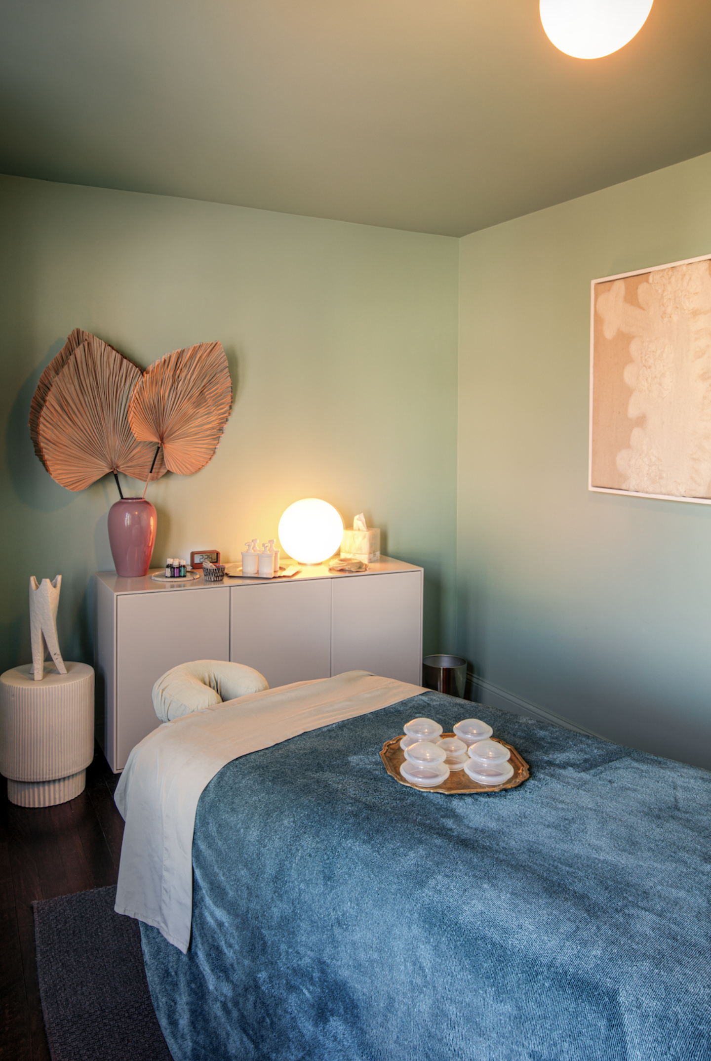Spa treatment room with teal walls, a massage bed covered in blue, and a glowing lamp atop a white cabinet.