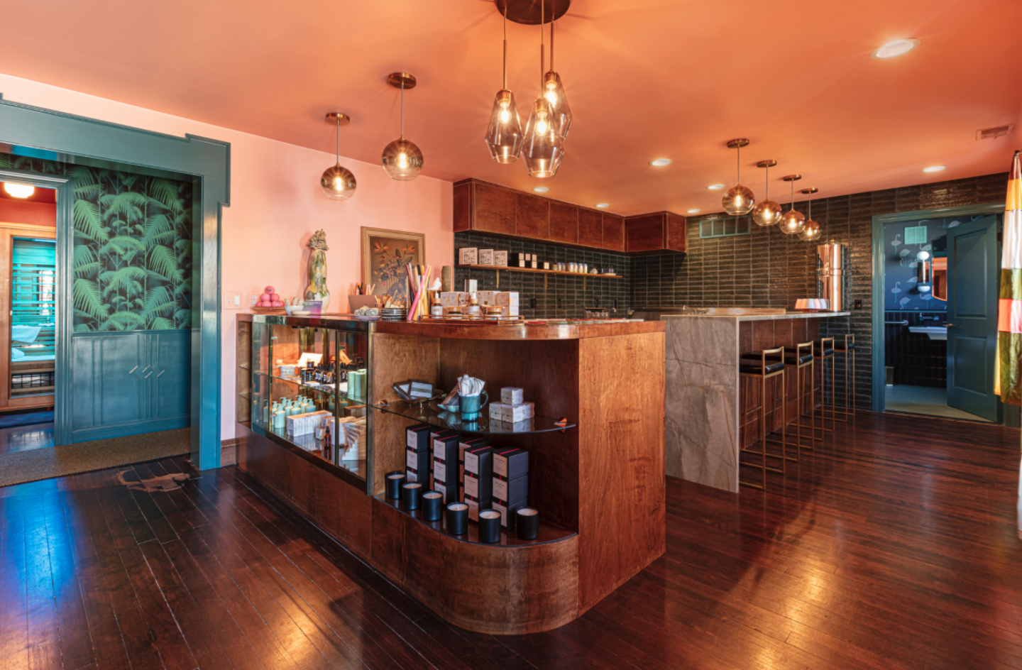 Warm, stylish café bar interior with a curved wooden counter, glass display case, and pendant lights; dark tiled back wall and wooden stools along the bar.