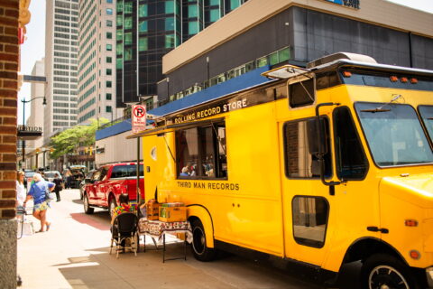 Yellow mobile record store bus on a city street, vinyl records displayed outside, pedestrians nearby.