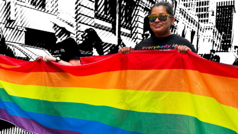 Two people hold a large rainbow Pride flag, one wearing sunglasses, in front of a black-and-white city mural.