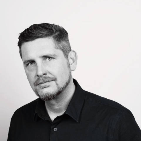 Portrait of a man with short dark hair and a light beard in a black collared shirt, against a plain light background.