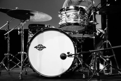 Close-up of a drum kit on a stage: bass drum with chrome hardware and a snare, cymbal to the left, microphone stands around.
