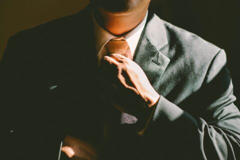 Close-up of a man in a dark suit adjusting a brown tie at the collar, partial face out of frame.