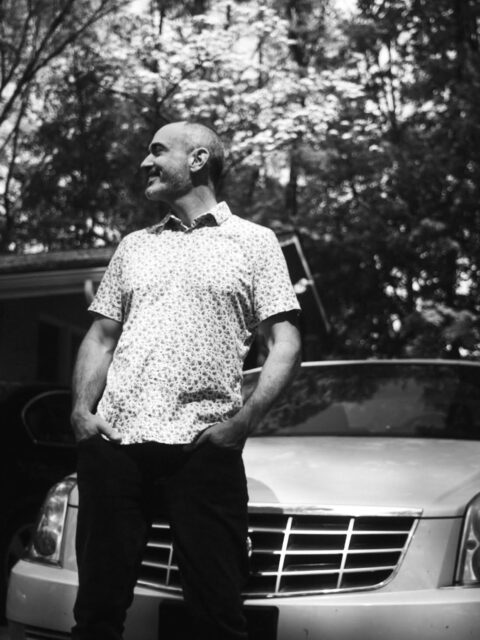 Man standing outside in a floral short-sleeve shirt, hands in pockets, smiling, with a car and trees in the background (black and white).