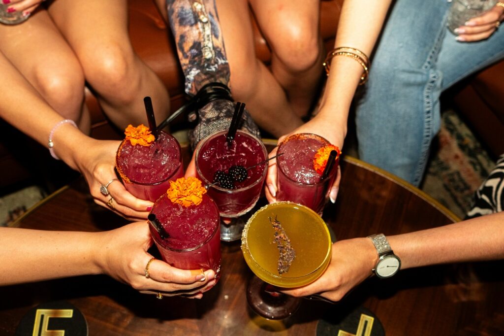 Group of friends clinking purple cocktails with orange flower garnishes on a wooden table in a bar room sense of celebration.
