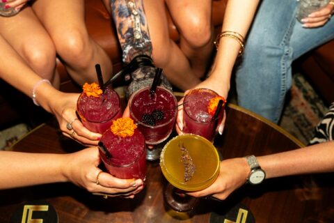 Group of friends clinking purple cocktails with orange flower garnishes on a wooden table in a bar room sense of celebration.