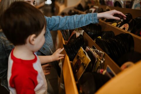 Child in a red-and-white shirt reaches into a crate of black vinyl records in a shop/market stall.