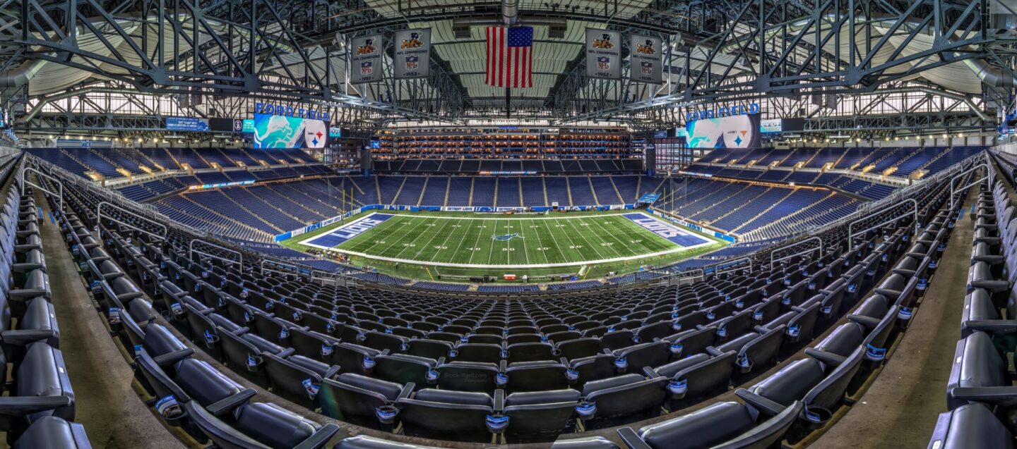 Panoramic view of Ford Field's interior with green football field, blue seating, and a hanging American flag above the arena.