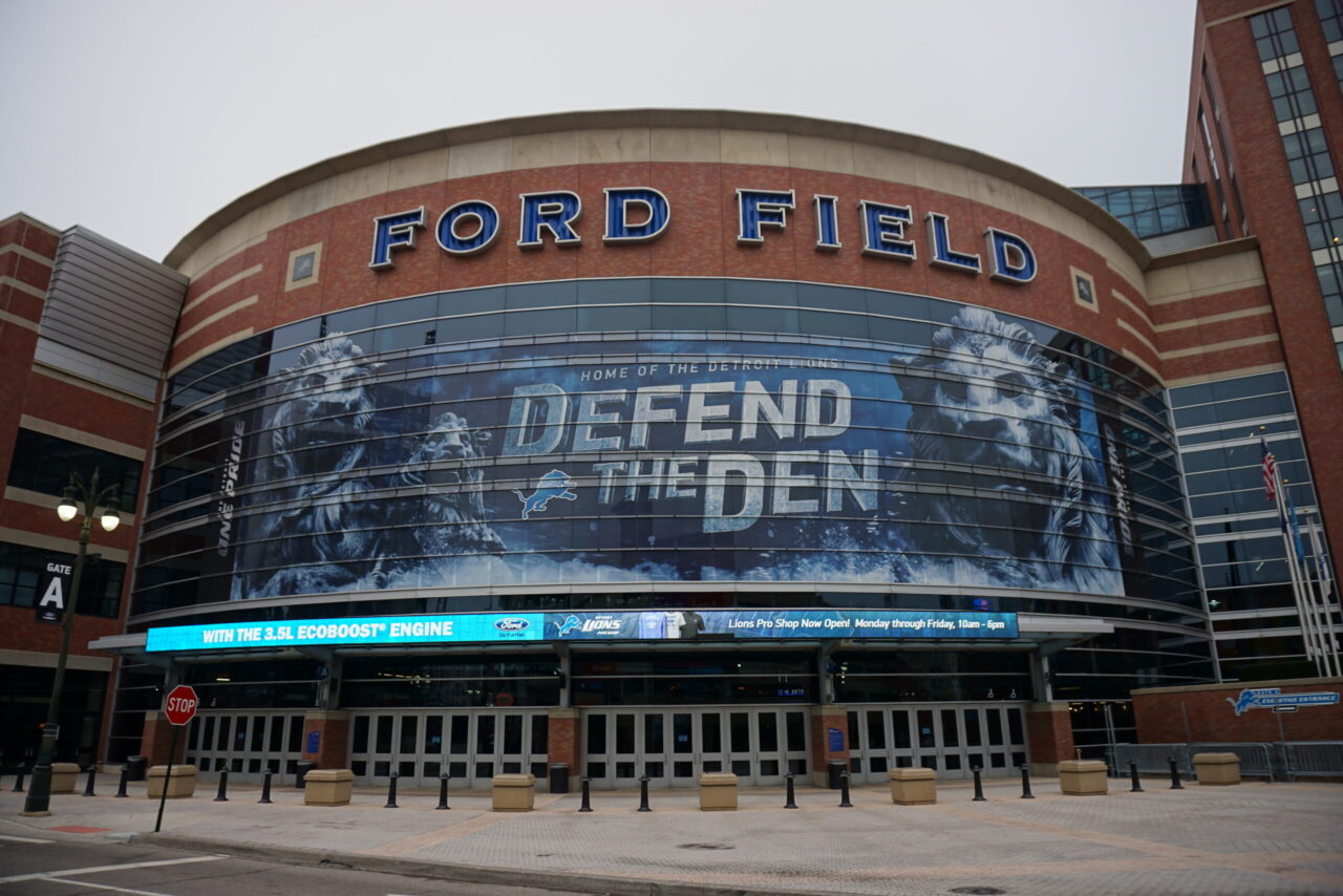 Exterior view of Ford Field stadium in Detroit with a large blue promotional banner reading 'DEFEND THE DEN' across the curved glass entrance façade.