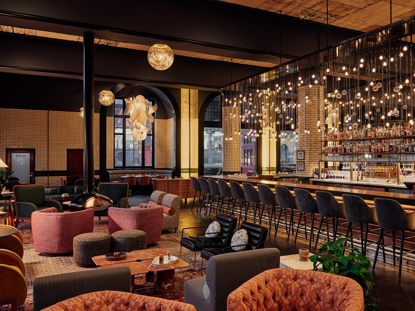 Upscale bar lounge with a long wooden bar, numerous pendant lights, and plush, colorful seating by warm brick walls.
