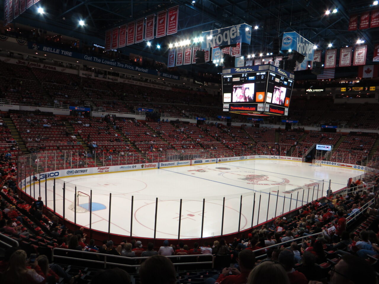 Ice hockey rink in a large arena with spectators in red seats and a central scoreboard overhead.