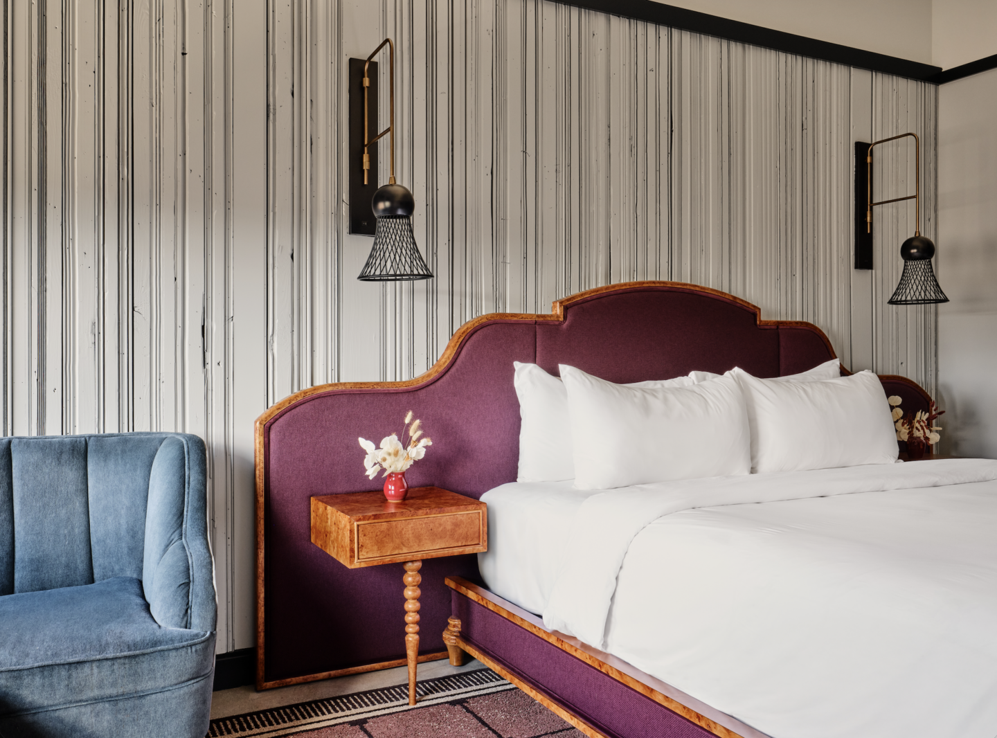 Hotel bedroom featuring a purple upholstered headboard, white pillows, and a pair of black wire shade lamps on striped gray walls.