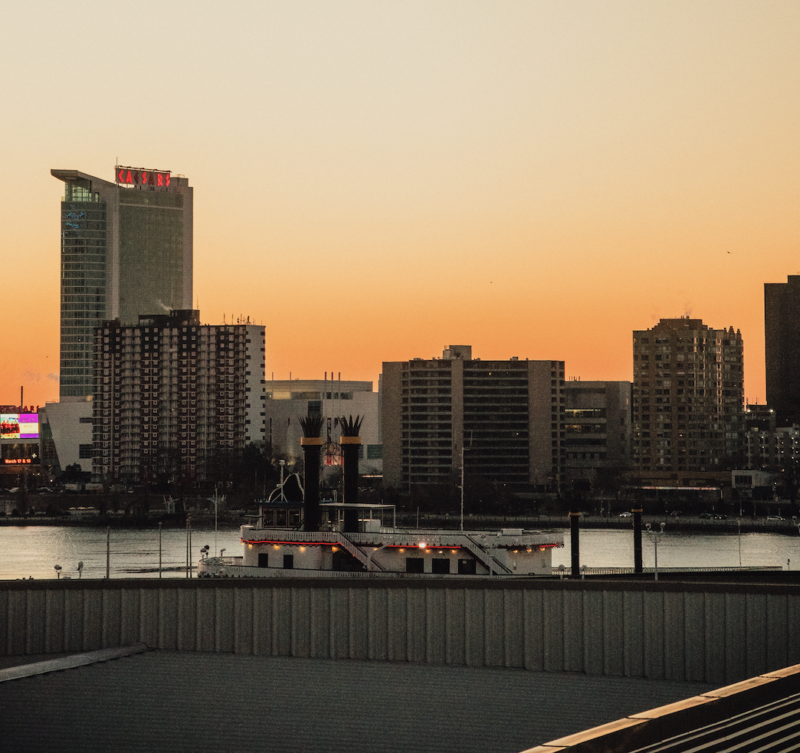 Sunset over a city skyline along a river, with a white riverboat in the foreground and tall buildings along the shore.