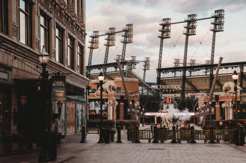 Street view toward Comerica Park with towering stadium floodlights and a historic building on the left near a pedestrian plaza.