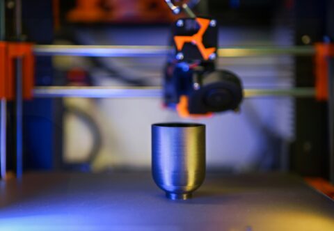 Close-up of a metallic cylindrical object on a 3D printer bed, with the printer head and orange components blurred in the background.