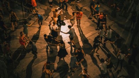 Overhead view of a crowd dancing in pairs on a stone plaza at night, long shadows stretching across the ground.