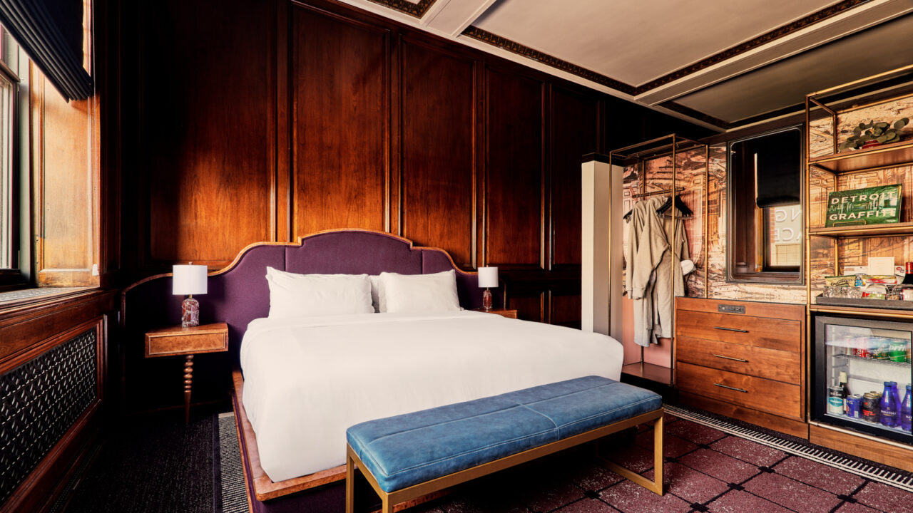 Hotel bedroom with white bed, purple upholstered headboard, dark wood wall panels, and a blue bench at the foot of the bed.
