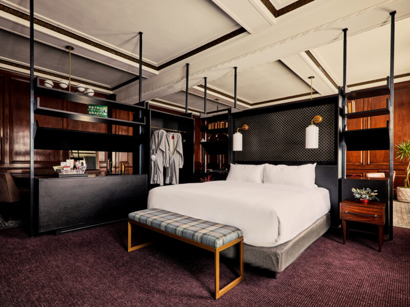 Luxurious hotel bedroom with a large white bed, dark wood panels, and an open wardrobe behind the headboard. A plaid bench sits at the foot of the bed.