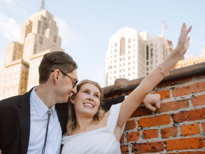 Bride in a white dress and groom in a suit lean against a brick wall in an urban rooftop setting, smiling at each other.