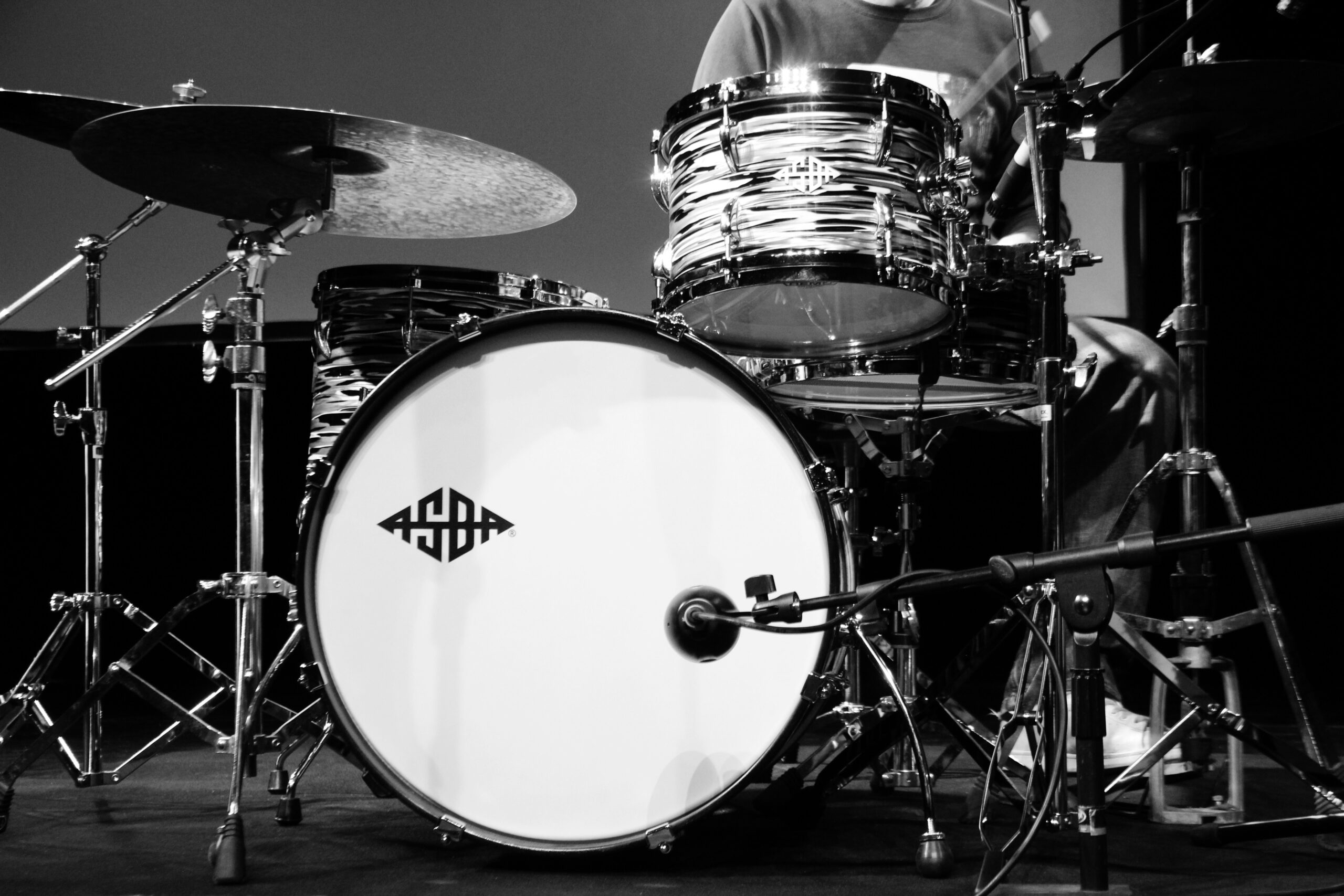 Black-and-white close-up of a drum kit on a stage, showing a large bass drum with logo, snare, toms, cymbals, and surrounding hardware.