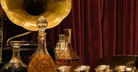 Vintage gramophone horn with crystal decanters and shot glasses on a dark bar against burgundy velvet curtains.