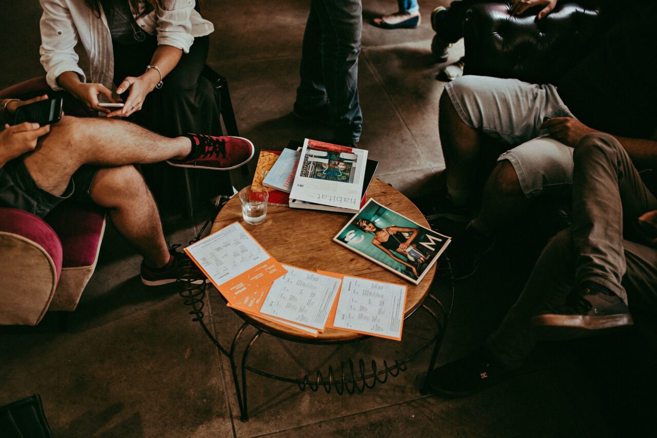 People seated around a round coffee table covered with magazines and orange flyers; their legs and feet are visible in a casual, indoor setting.