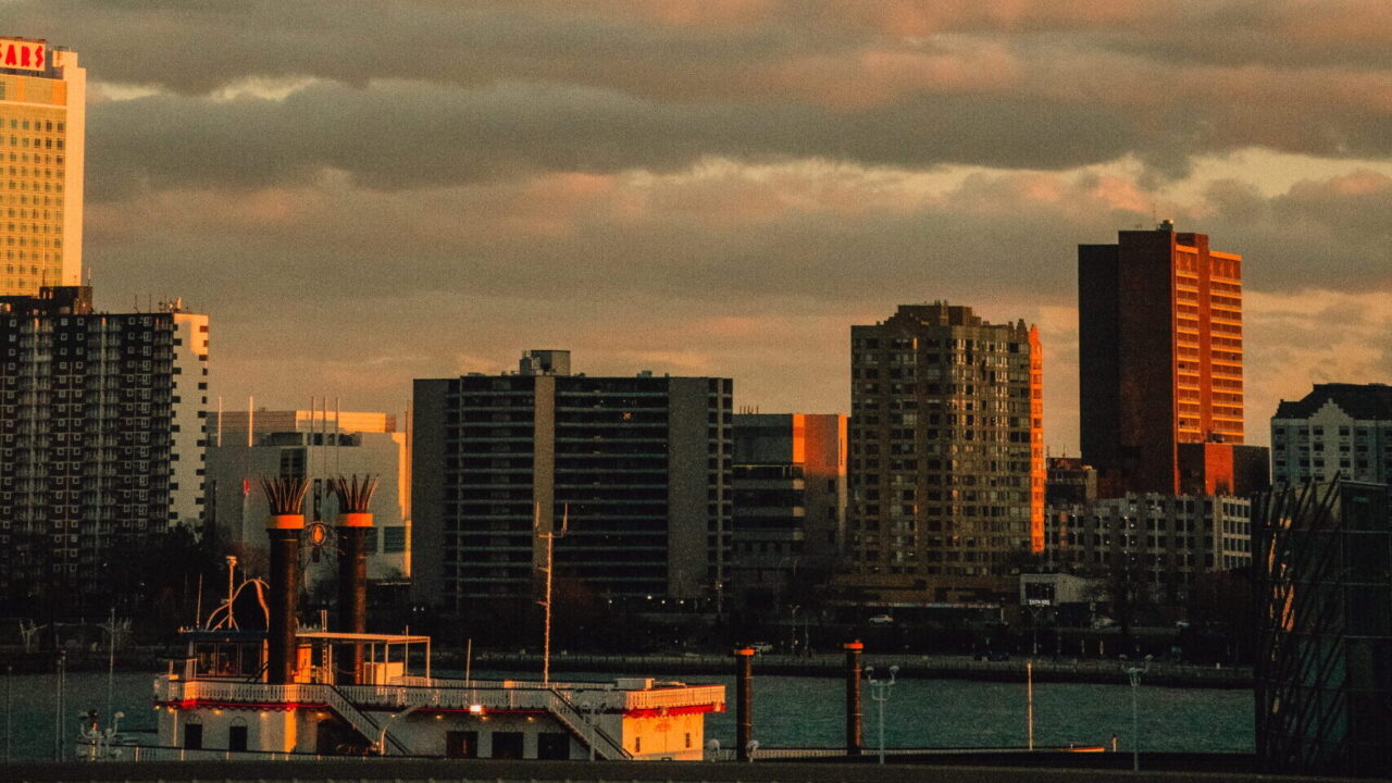 City skyline at sunset with dark clouds and orange-pink sky over river, silhouettes of tall buildings along the water's edge and docked ships in the foreground.