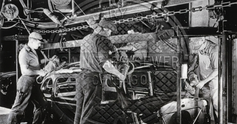 Black-and-white photo of factory workers assembling a car on an old assembly line, handling parts and tools.