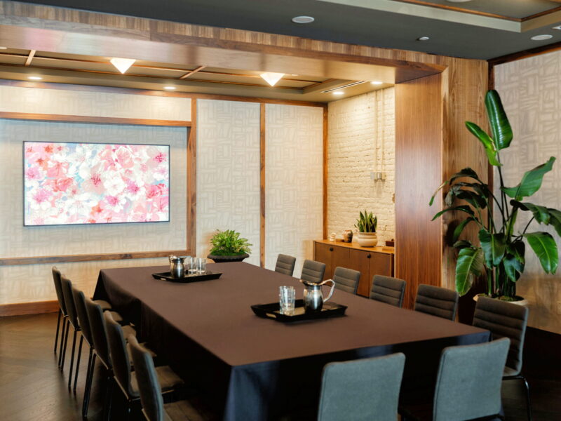 Conference room with a long black-table setup, gray chairs, plants, and a pink floral painting on the wall behind.