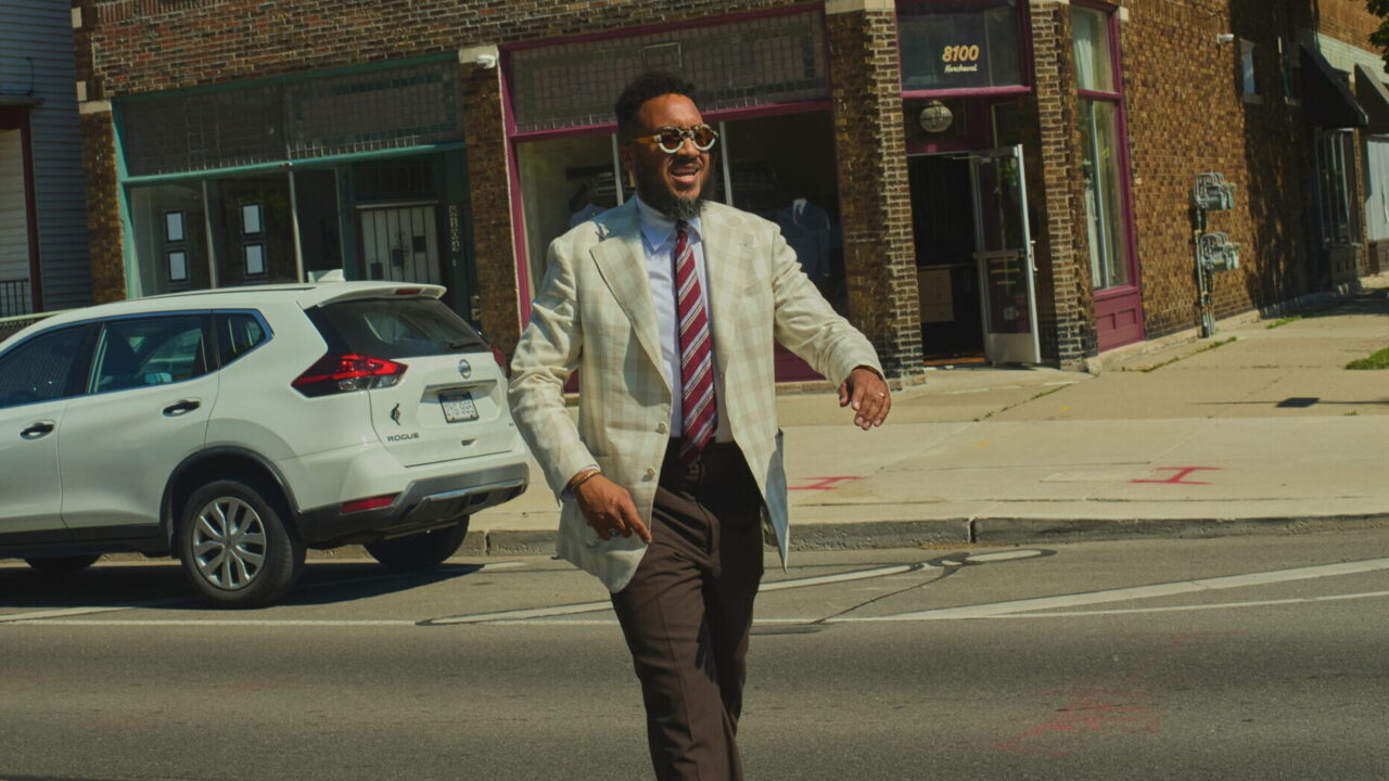 Man in a light blazer and sunglasses walking across a city street with brick storefronts and blue sky behind him.
