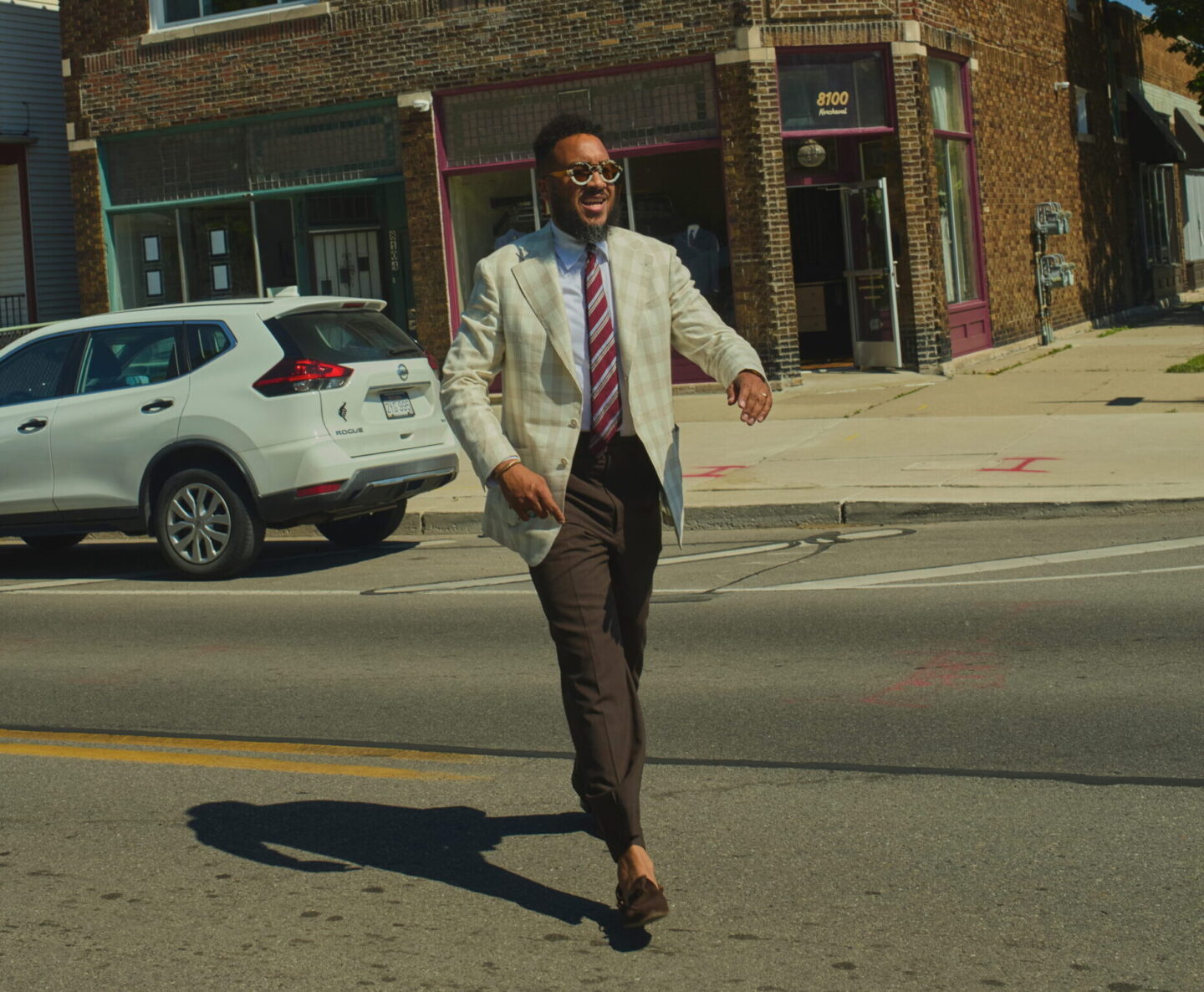 Man in a light blazer and sunglasses walking across a city street with brick storefronts and blue sky behind him.