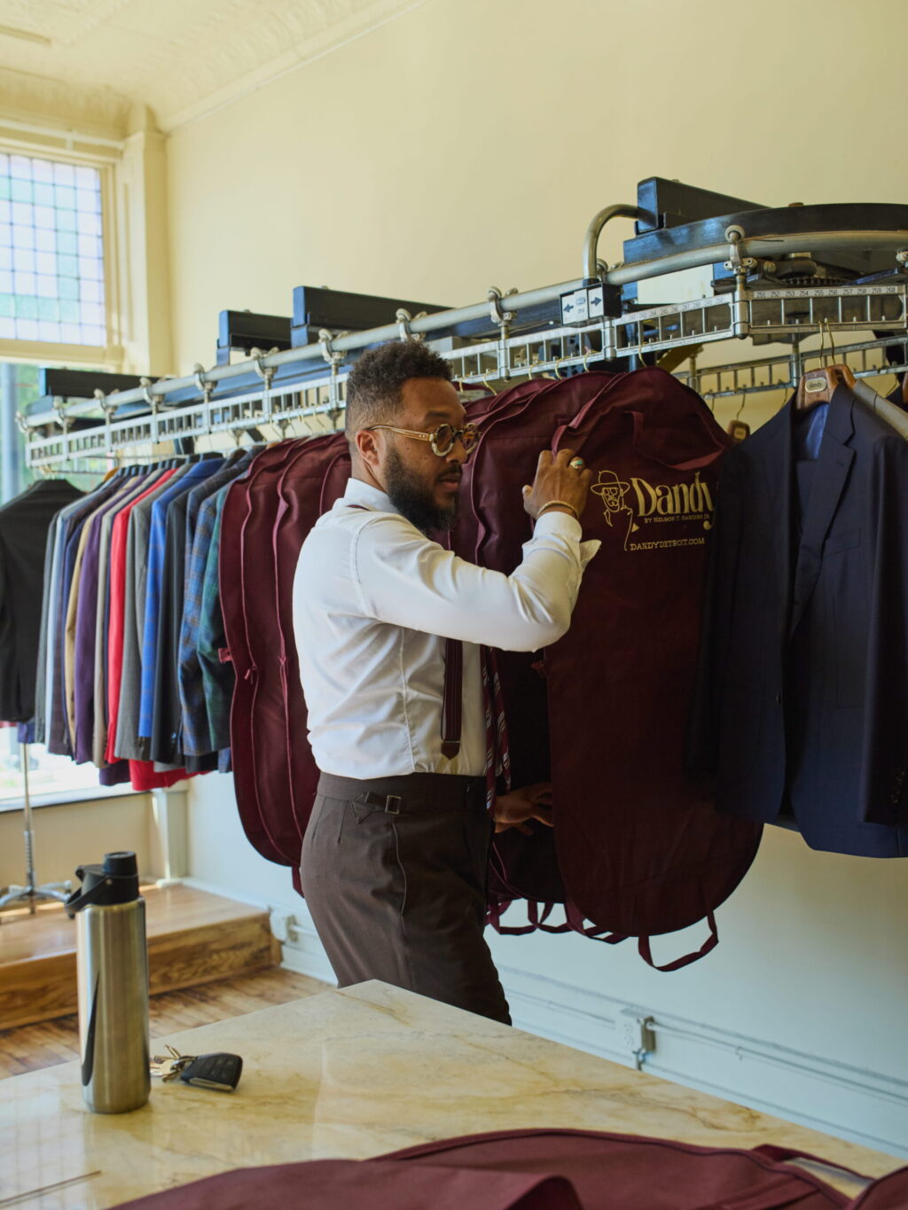 Man in a white shirt and suspenders examines a burgundy suit jacket on a rack in a tailoring shop, with colorful jackets lined behind him.