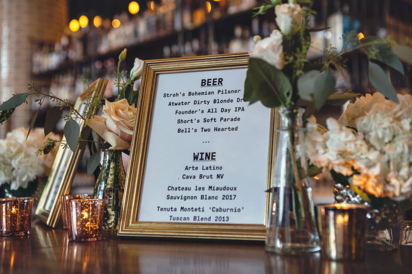 Framed white menu board on a bar with flowers and candles, listing beer and wine options.