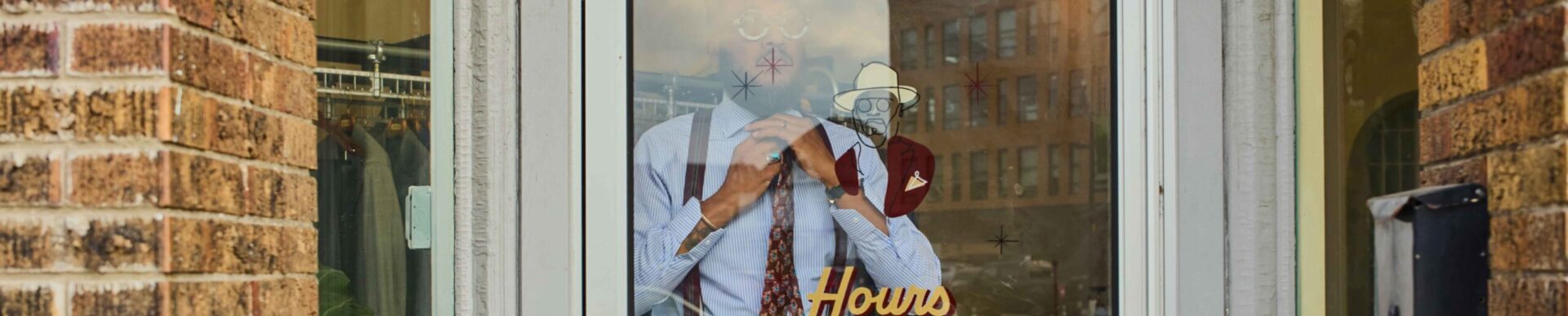 Storefront window display of a man in a blue shirt and tie adjusting his collar; gold text reads 'Hours Tue-Fri 11-6'.