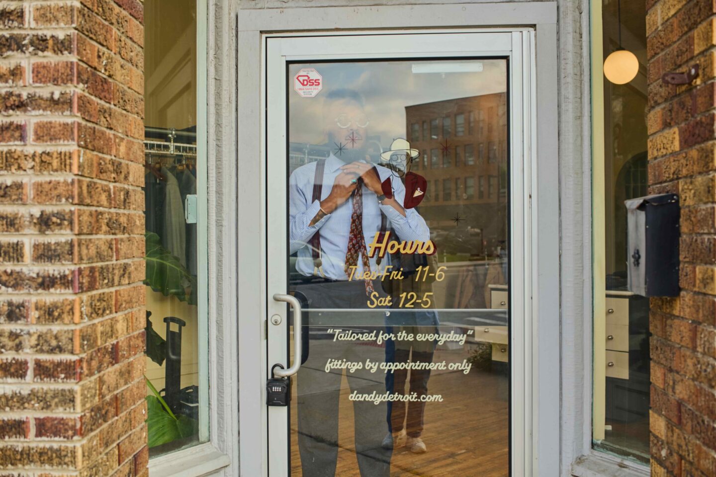 Glass storefront door with hours listed and cartoon drawings; reflection of a man in a shirt and tie seen in the glass.