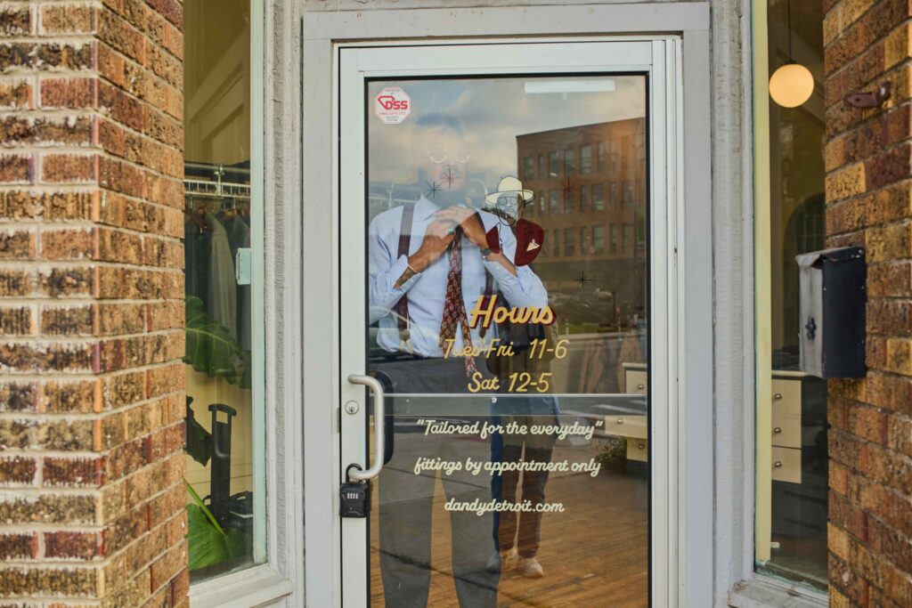 Storefront glass door with a brick exterior; reflection of a man in a shirt and tie with suspenders in the door’s glass, plus decorative drawings and yellow hours text visible on the glass.