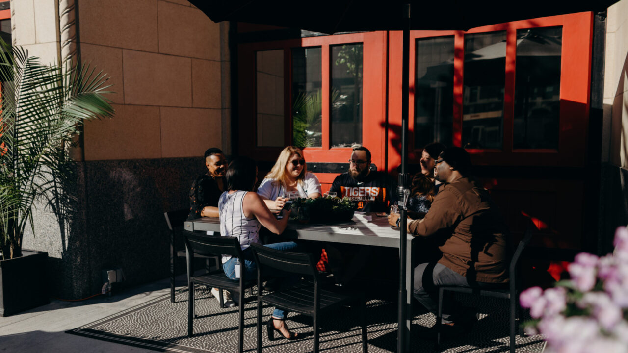 Group of friends seated around a rectangular outdoor table, talking and smiling in sunlight, with a red storefront door behind them.