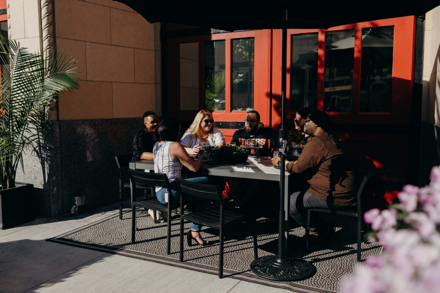Group of friends seated around a rectangular outdoor table, talking and smiling in sunlight, with a red storefront door behind them.