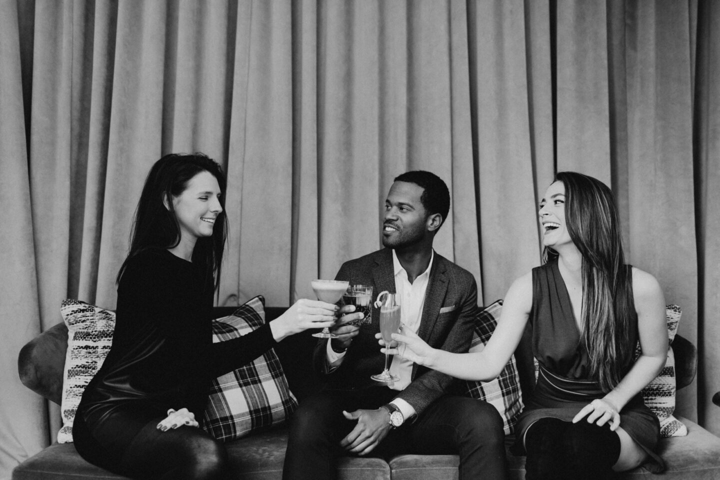 Four friends on a sofa toasting with drinks, smiling and chatting together at a casual gathering.
